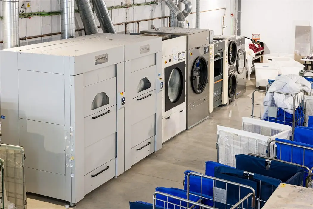 Industrial washing machines and dryers in a row next to linen trolleys at the plant