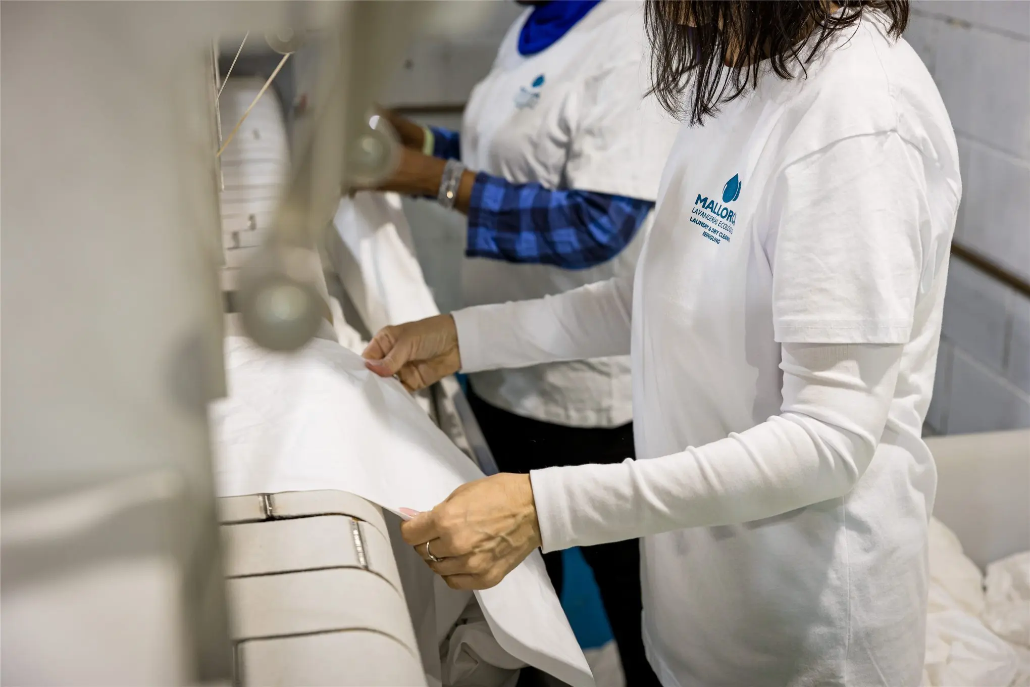 Two Lavandería Mallorca workers smoothing white table linen on the ironer